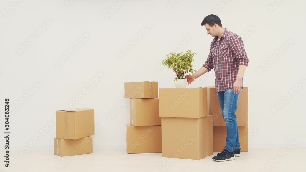 Handsome young man appearing with flowerpot, putting it on the pile of boxes, hands into pockets, looking around new apartments. Handsome young man appearing with flowerpot, putting it on the pile of boxes, hands into pockets, looking around new apartments.