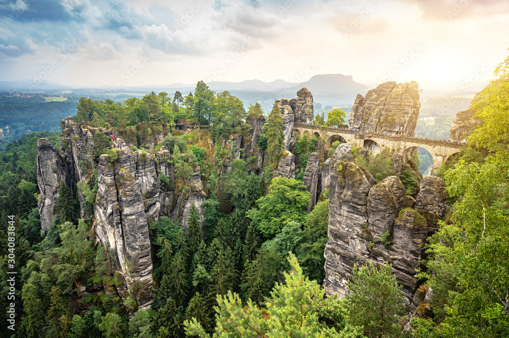 Beautiful panoramic view of famous Bastei Bridge with Elbe Sandstone mountains in Saxon Switzerland National Park with dark clouds in golden morning light at sunrise in spring, Saxony, Germany