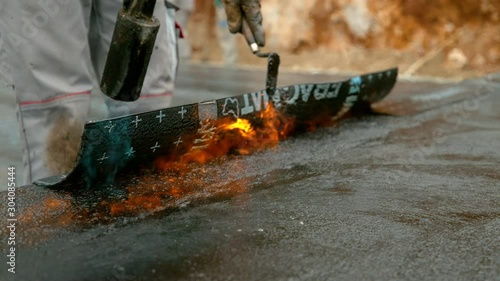 SLOW MOTION, CLOSE UP: Unrecognizable laborer lays black tar insulation over the building's foundation. Man runs a fire extinguisher along a sheet of bitumen coating being melted onto the ground. photo