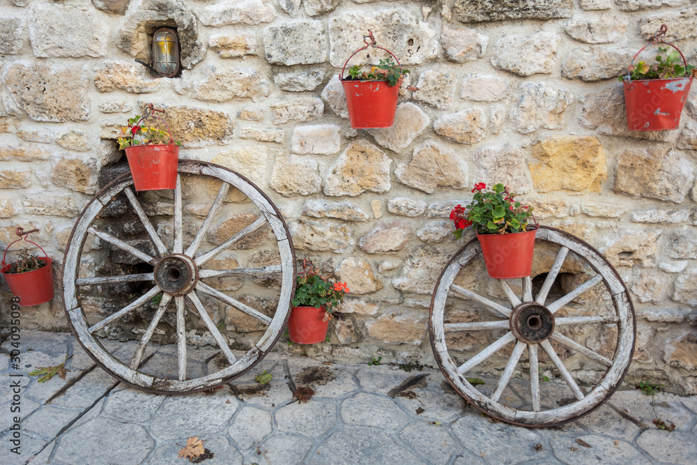 Unusual street decor in Greece. Two old vintage wooden wheels standing near stony house wall decorated with many buckets with growing flowers. Horizontal color photography.