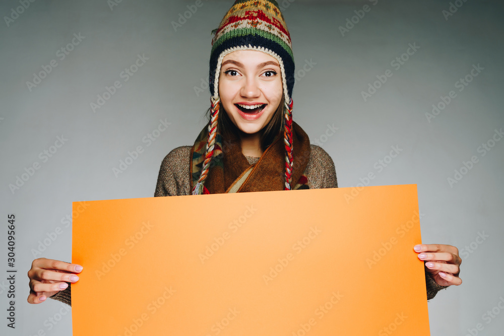 a girl in a winter hat opens her mouth in surprise looking joyfully at the camera holding a yellow sheet of paper in her hands. gray background a girl in a winter hat opens her mouth in surprise looking joyfully at the camera holding a yellow sheet of paper in her hands. gray background