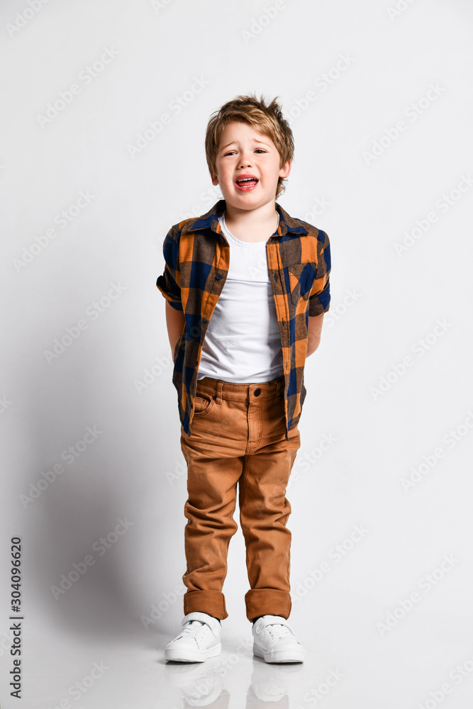 Little upset kid boy in plaid shirt is holding his hands behind his back and crying loud like he got lost on white Little upset kid boy in plaid shirt is holding his hands behind his back and crying loud like he got lost on white