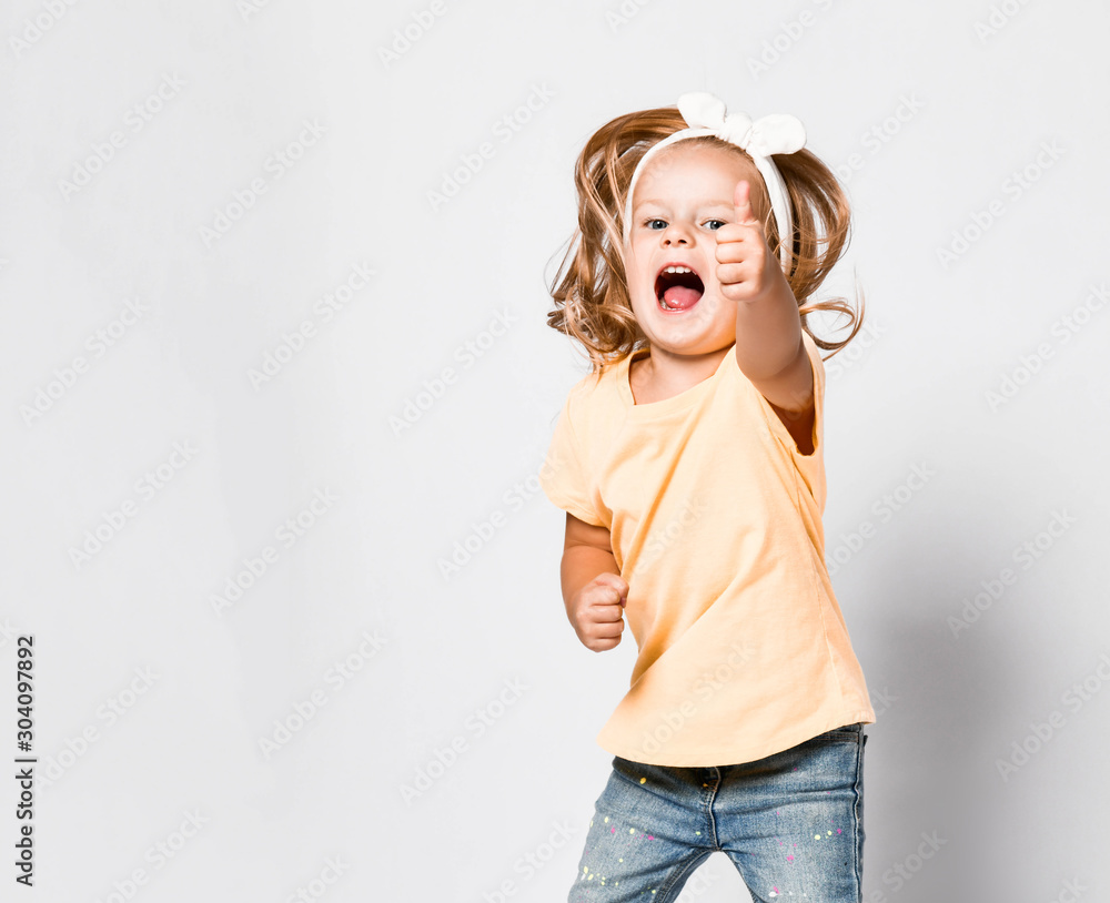 Happy screaming baby girl in yellow t-shirt is having fun good time, jumping, playing, showing thumb up sign, good job Happy screaming baby girl in yellow t-shirt is having fun good time, jumping, playing, showing thumb up sign, good job