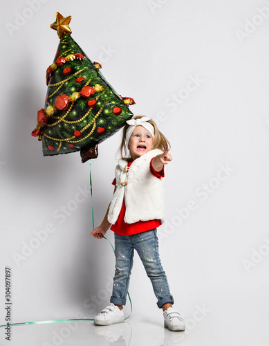 Screaming frolic baby girl in red t-shirt and white waistcoat with Christmas tree balloon is pointing at free copy space
