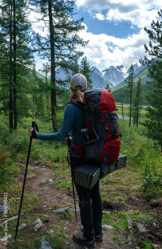 Girl tourist with backpack and poles looking at the beautiful summer landscape with mountain peaks. Hiking, the concept of research, travel and expeditions.