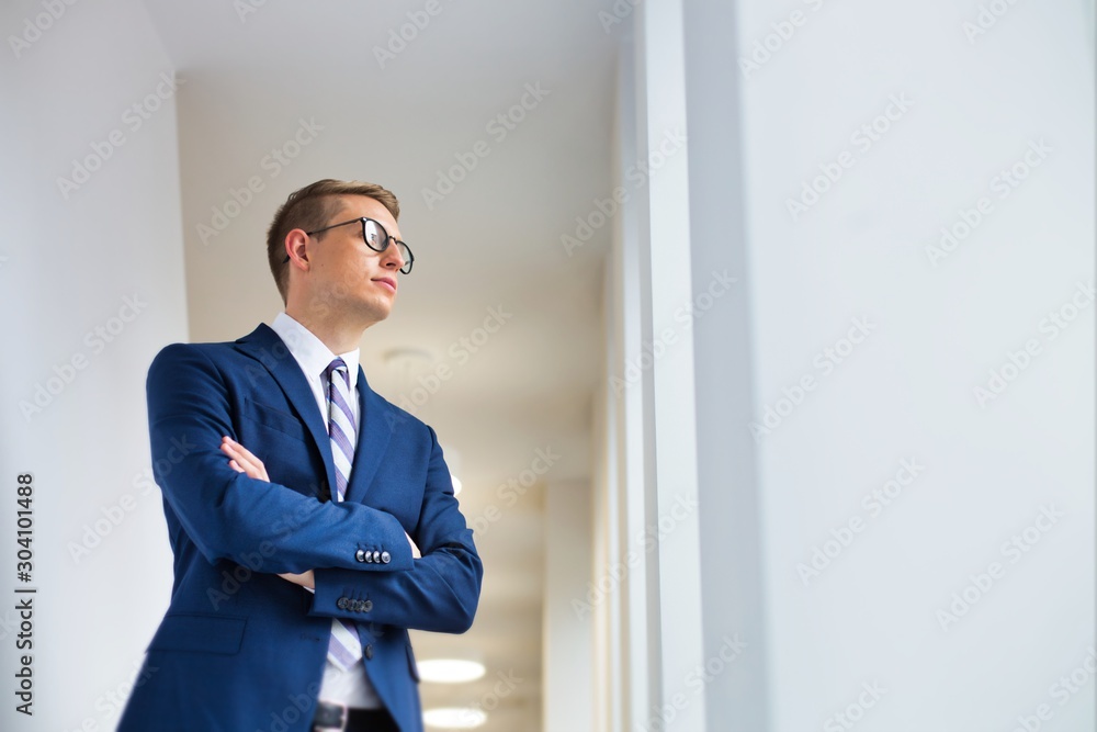 Young thoughtful businessman standing with arms crossed looking away in office corridor Young thoughtful businessman standing with arms crossed looking away in office corridor