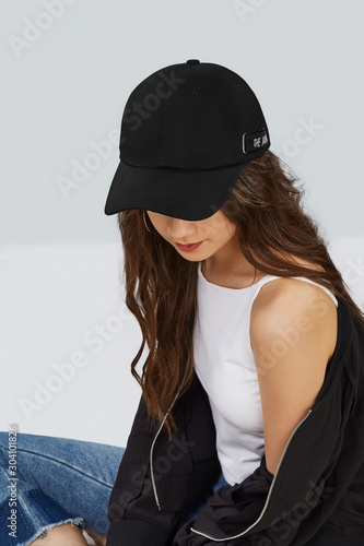 Cropped upward shot of a dark-haired girl, wearing black baseball cap, white tank top, jeans with scuffs and black jacket. The cap peak is closing upper part of a face. 