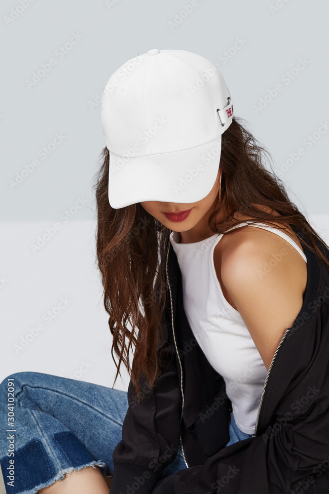 Cropped upward shot of a dark haired girl wearing white baseball cap white tank top jeans with scuffs and black jacket. The cap peak is closing upper part of a face. Stock Photo