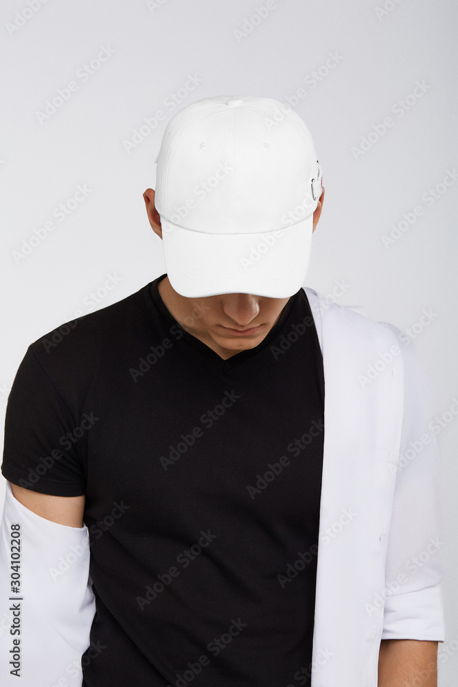 Cropped upward shot of a dark-haired man, wearing white baseball cap, black t-shirt and white shirt. The cap peak is closing upper part of a face. He is posing on grey background. Cropped upward shot of a dark-haired man, wearing white baseball cap, black t-shirt and white shirt. The cap peak is closing upper part of a face. He is posing on grey background.