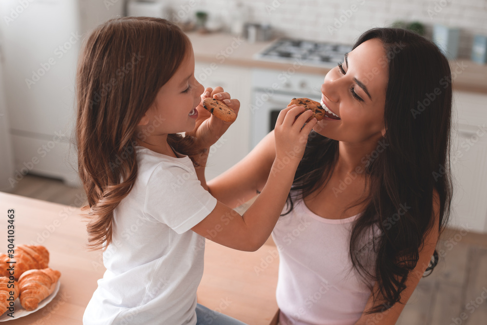 portrait of mother and daughter feeding each other with biscuits on the kitchen - obrazy, fototapety, plakaty 