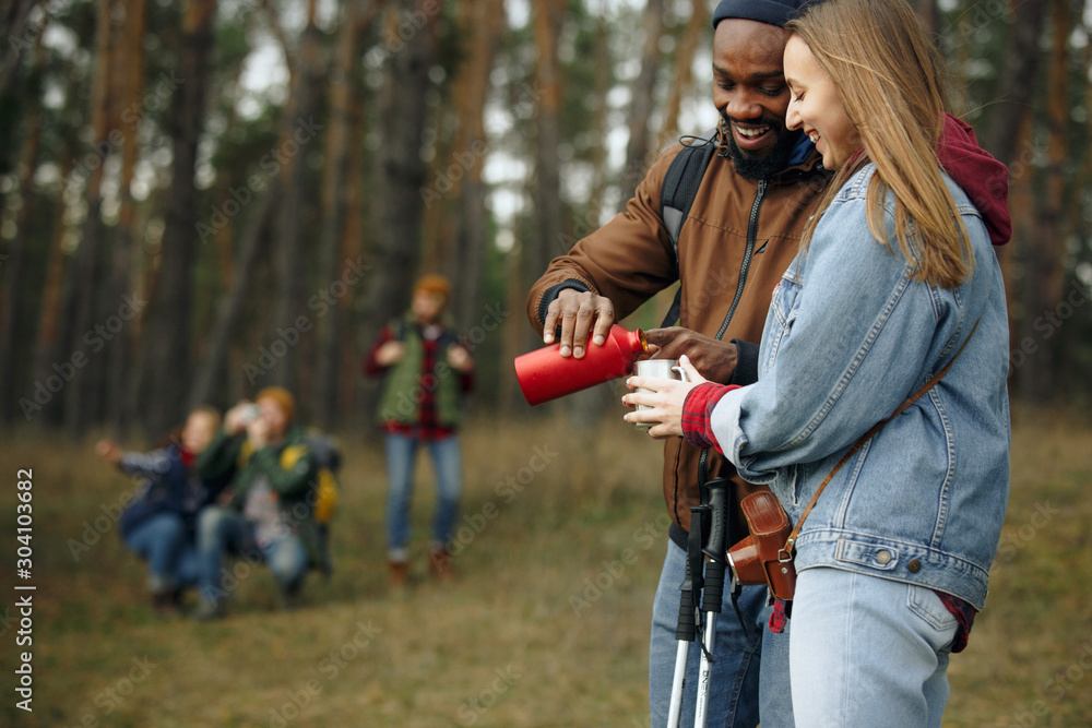 Obraz premium Group of friends on a camping or hiking trip in autumn day. Men and women with touristic backpacks in the forest, talking, laughting. Leisure activity, friendship, weekend. Drink tea or coffee.