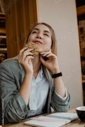 Lifestyle Portrait of smiling young casual blonde woman eating a cookie and drinking coffee  tea in cafe. Happy girl eating a dietetic gluten free cookie