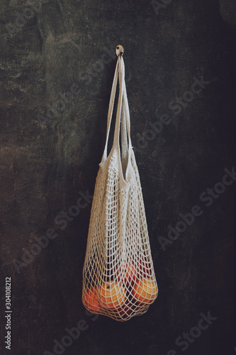 Reusable Mesh cottone Bags hanging on the wall in house. Eco friendly package for grocery with fruits on dark background. Reusable life, Zero waste, no plastic concept with copy space photo