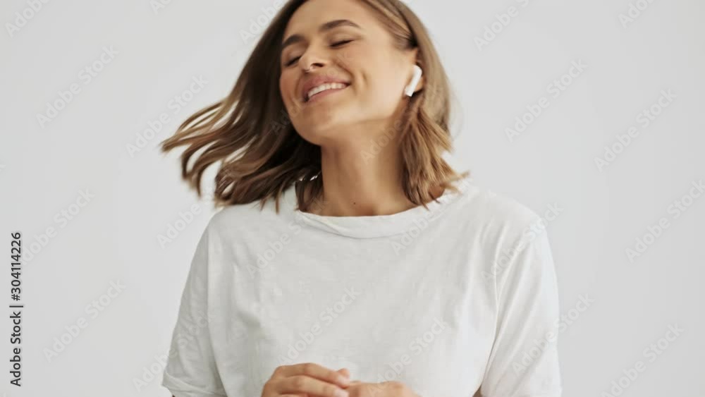 Relaxed smiling young woman wearing a white basic t-shirt listening to music on her wireless headphones with her eyes closed while swinging from one side to another isolated over white background Relaxed smiling young woman wearing a white basic t-shirt listening to music on her wireless headphones with her eyes closed while swinging from one side to another isolated over white background