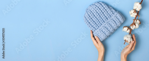 Flat lay fashionable blue knitted winter hat in female hands cotton flowers on blue background top view. Stylish woolen hat concept of winter accessories for the cold. Advertising shopping winter sale