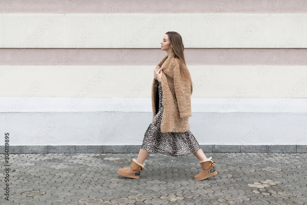 artificial fur coat, long leopard print dress, ugg boots, a small shoulder  bag, a beautiful white model girl with long hair poses cheerfully walks on  the street near the wall Stock Photo