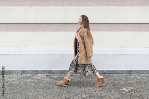 artificial fur coat, long leopard print dress, ugg boots, a small shoulder bag, a beautiful white model girl with long hair poses cheerfully walks on the street near the wall