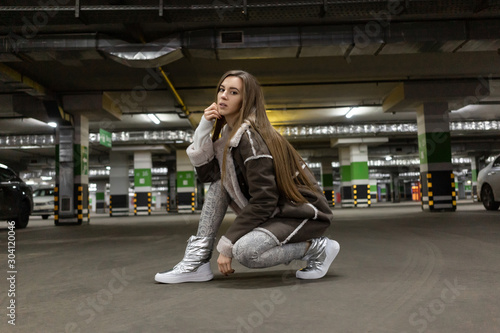 leather sports shoes white gray demi season. Beautiful European model with long hair squats in an underground parking lot. Sheepskin coat. Snake skin print leggings photo