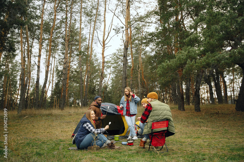 Group of friends on a camping or hiking trip in autumn day. Men and women with touristic backpacks having break in the forest, talking, laughting. Leisure activity, friendship, weekend. Eat and drink.