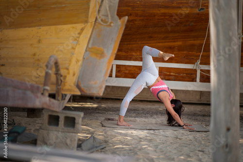 Beautiful brunette fitness model and yogi wearing a pink crop top and gray yoga tights performs a yoga pose in a rustic boatyard 