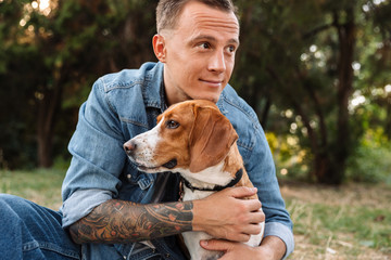 Photo of beautiful young man smiling and hugging his canine dog in park