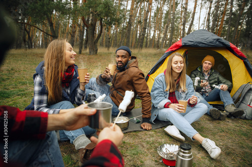 Group of friends on a camping or hiking trip in autumn day. Men and women with touristic backpacks having break in the forest, talking, laughting. Leisure activity, friendship, weekend. Eat and drink. © master1305