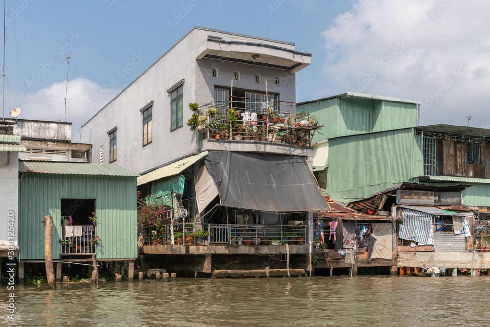 Cai Be, Mekong Delta, Vietnam - March 13, 2019: Along Kinh 28 canal. Rather nice green and gray dwellings built half over the brown water under blue cloudscape. 