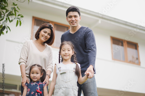 Young Asian family love, father mother and two daughter standing at outdoor in front of home. girl and parent looking at camera which smiling and felling happy and background is a white house.