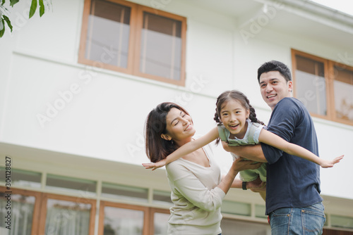 Young Asian family love, father mother and daughter standing playing and fun at outdoor in front of home. girl and parent looking at camera which smiling and felling happy. background is white house.