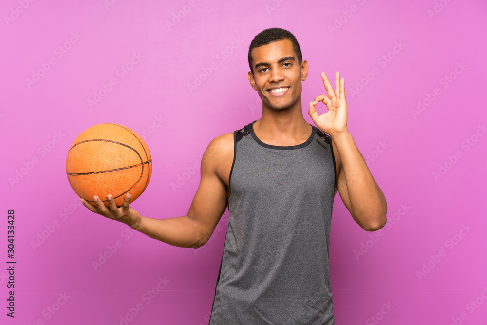 Young sport man with ball of basketball over isolated purple wall showing ok sign with fingers