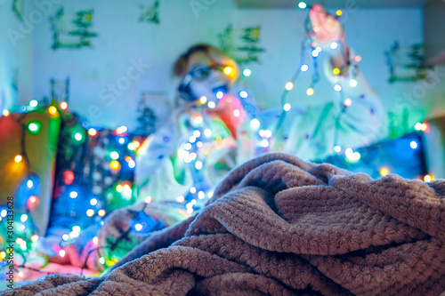 winter holidays preparing cozy domestic atmosphere lonely girl unfocused silhouette background happy emotion with garland unfocused colorful light illumination, blanket texture foreground 