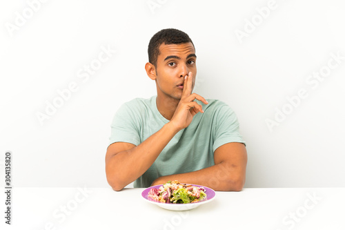 Young handsome man with salad in a table doing silence gesture
