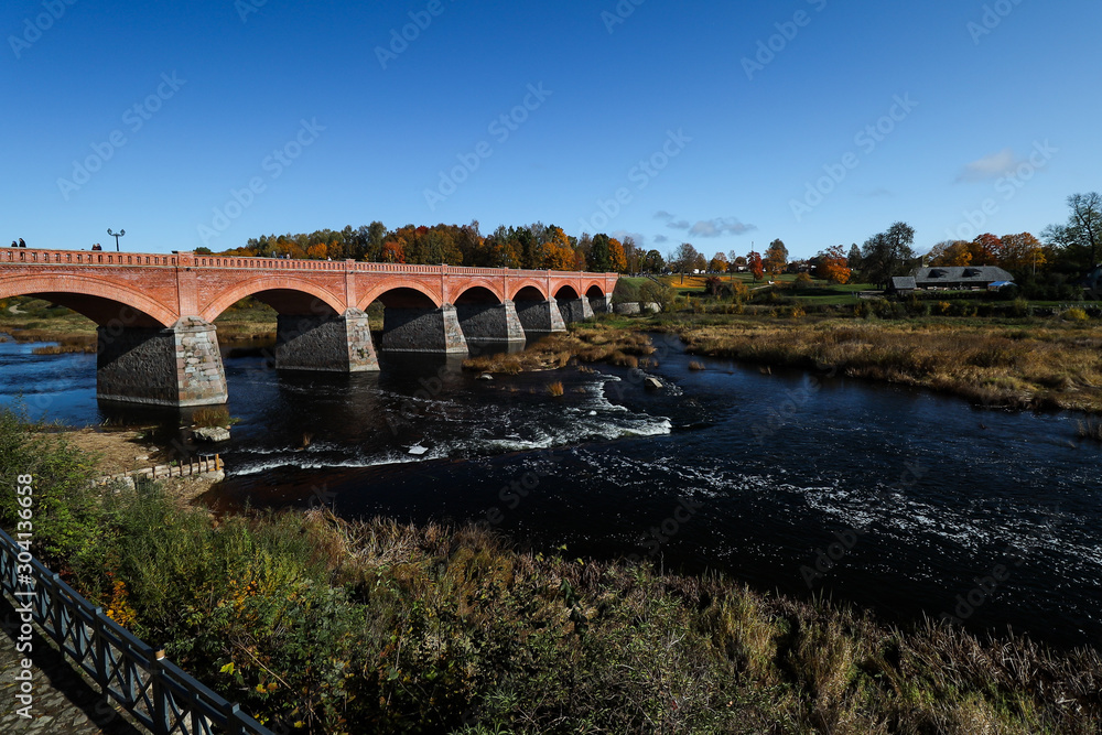 Fototapeta premium Daytime view of historic red brick bridge over river Venta in Europe-Latvia-Kuldīga.
