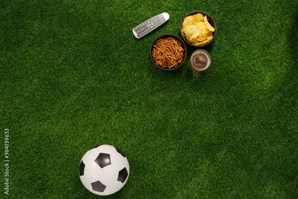 Naklejka premium Soccer ball on a green field and ottoman for a fan with snacks and a TV remote control. flat lay.Copy space.