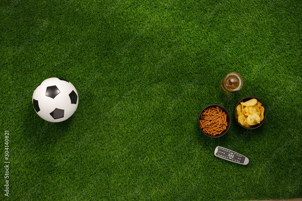 Naklejka premium Soccer ball on a green field and ottoman for a fan with snacks and a TV remote control. flat lay.Copy space.