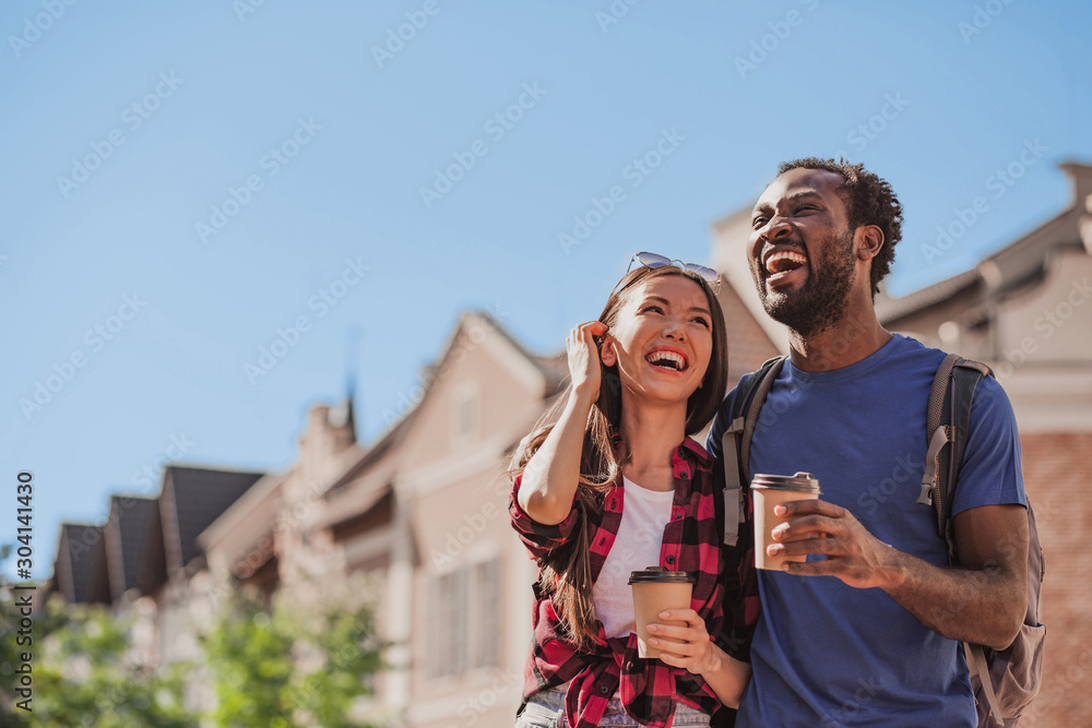 Naklejka premium portrait of laughing asian girlfriend and african american boyfriend walking in the city with coffee cups