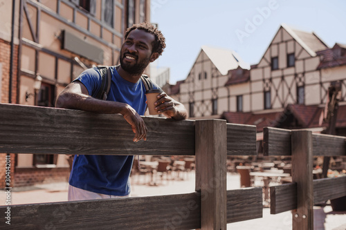 smiling young man with backpack and coffee cup standing at wooden fence © Afshar Tetyana