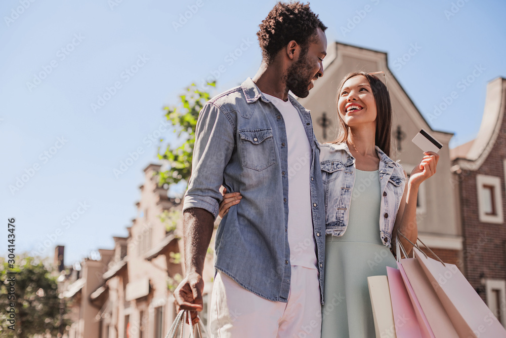 cheerful man and woman looking at each other while walking on the street with shopping bags
