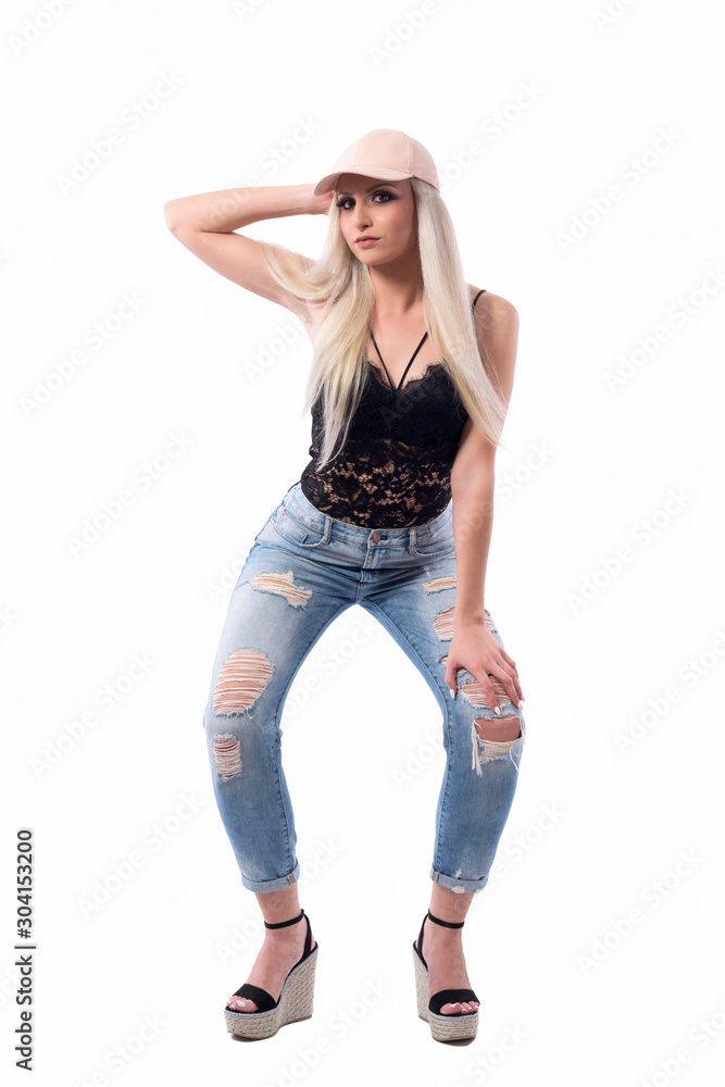 Modern trendy style blond woman wearing cap and jeans posing and looking at camera with hand on knee. Full body isolated on white background. Modern trendy style blond woman wearing cap and jeans posing and looking at camera with hand on knee. Full body isolated on white background.