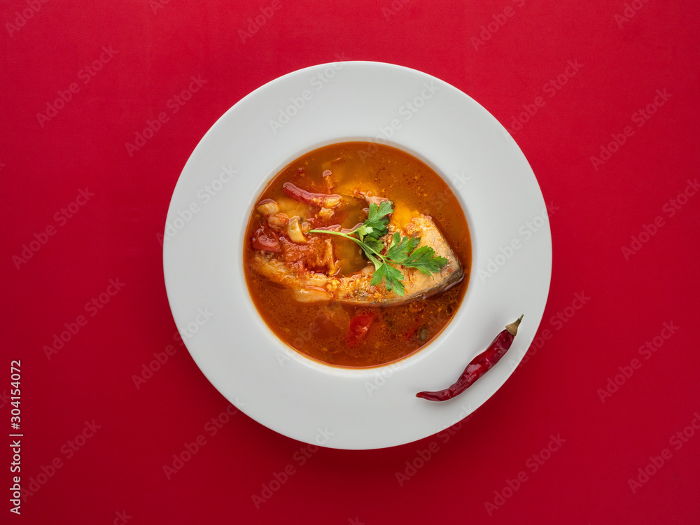 Fish soup in white plate with chilli pepper on red background. Overhead view. Fish soup in white plate with chilli pepper on red background. Overhead view.