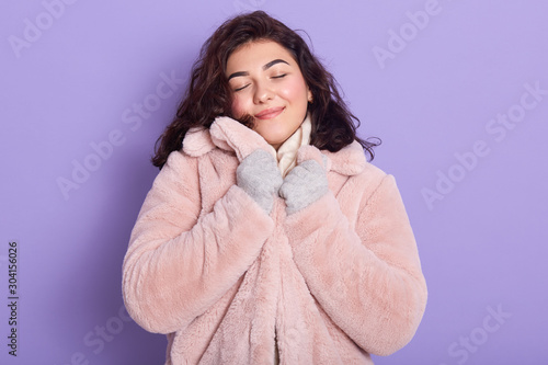 Beautiful girl wearing pale pink faux fur coat, scarf and gloves, adorable female looks pleased and satisfied, keeps hands on her collar, posing with closed eyes isolated over lilac studio background. photo