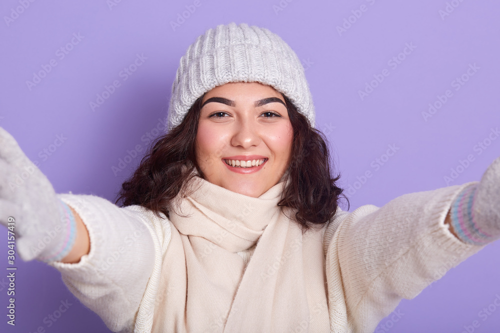 Portrait of good looking young lady extending her hands, smiling sincerely, looking directly at camera, wearing white hat, scarf, gloves and sweater, being in high spirits. People and winter concept. Portrait of good looking young lady extending her hands, smiling sincerely, looking directly at camera, wearing white hat, scarf, gloves and sweater, being in high spirits. People and winter concept.