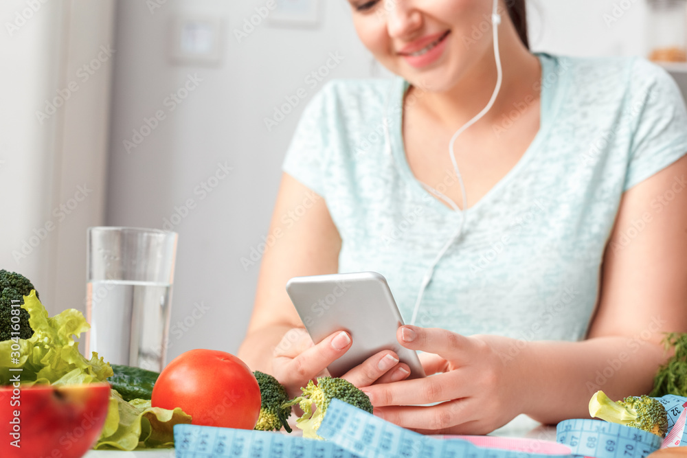 Body Care. Chubby girl in earphones sitting at kitchen table with smartphone listening music close-up joyful blurred background