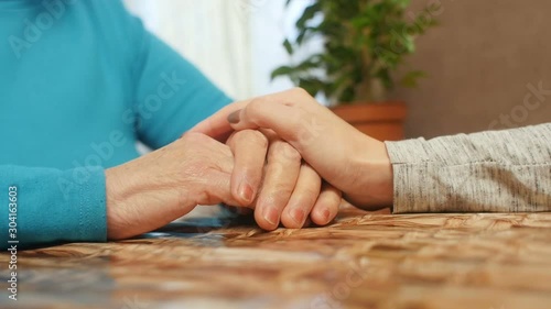 wrinkled hands of an elderly worried woman on kitchen table, granddaughter consoles beloved grandmother holding her hands, concept care and family relationship slow motion