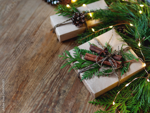 Christmas presents wrapped in craft paper with natural fir tree and thuja branches as decoration. Wooden table with hand made New Year gifts and light bulbs.