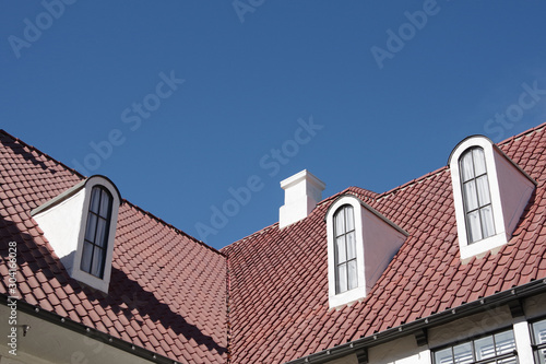 Low angle view of an angled red tile roof with tall slim dormer windows under a bright blue sky