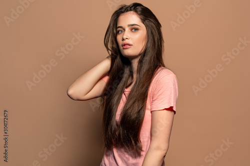 Freestyle. Young girl in t-shirt standing studio isolated on brown touching hair looking camera curious