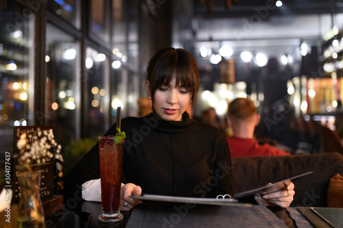 Beautiful young caucasian woman sitting in restaurant and attentively looking at menu choosing what to order. Dinner outdoor. Visiting cafe in evening after job.