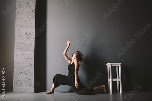 Fototapeta Naklejka Na Ścianę i Meble -  social theme of female loneliness is pain suffering. Abastration of male domestic violence. Young beautiful Caucasian woman in black clothes, strange pose, white chair big dark room. Fear and despair