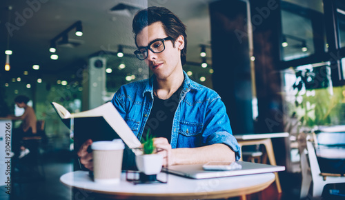 Thoughtful hipster guy in optical spectacles reading knowledge literature in coffee shop for making research of information for course work, pondering man holding textbook for education in hands